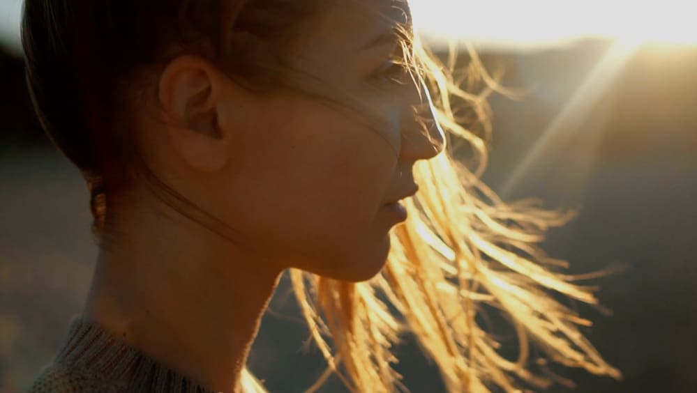 Rosto de uma mulher em perfil laterial com vento batendo em seus cabelos com a luz do sol a iluminando